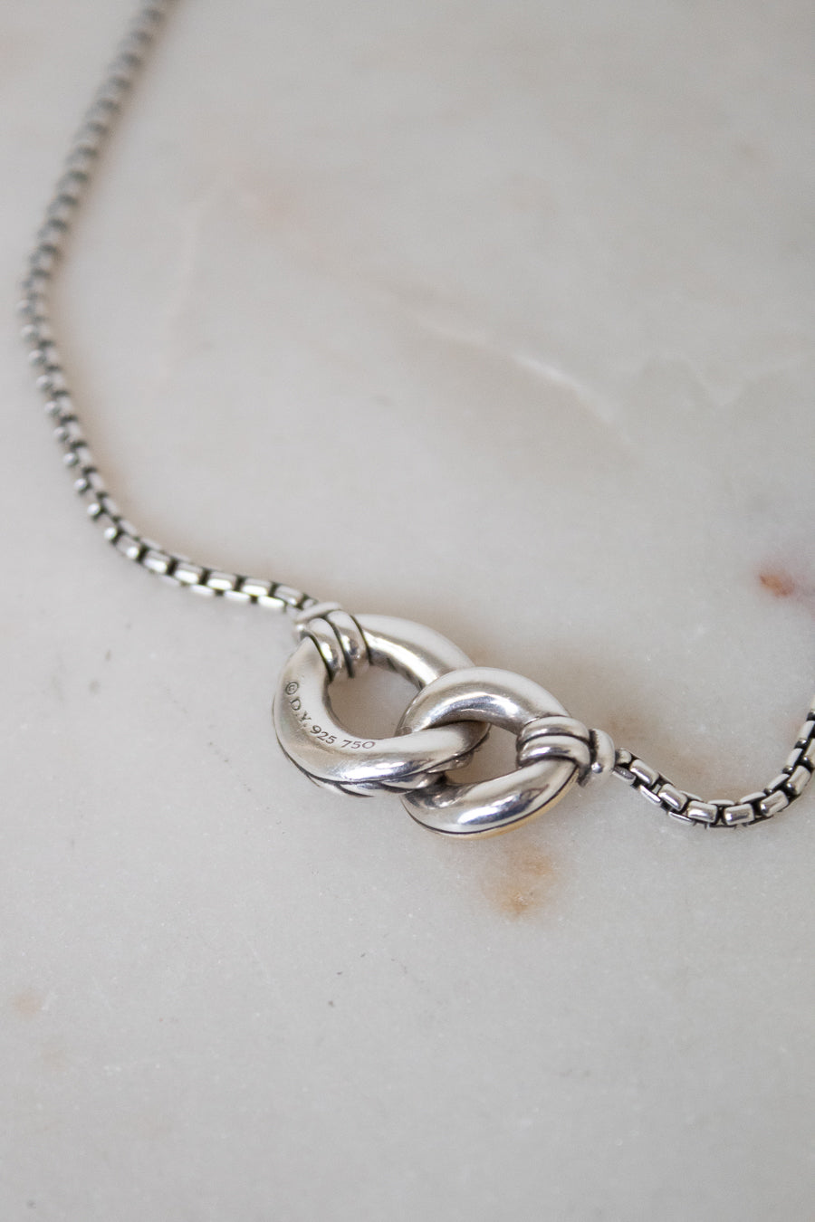 A close-up of a silver box chain necklace showing the back of an interlocked ring pendant, resting on a light beige marble surface.