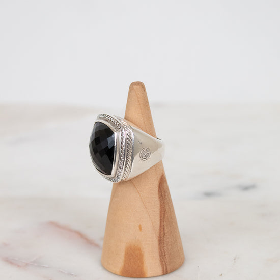 A silver ring with a large black stone, displayed on a small wooden cone stand against a light gray background.