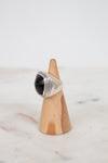 A silver ring with a large black stone, displayed on a small wooden cone stand against a light gray background.
