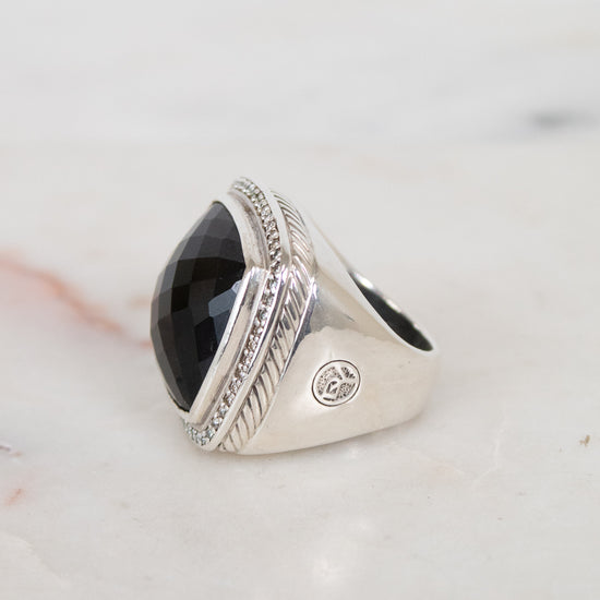 A silver signet ring with a large, faceted black oval stone, resting on a light marble surface.