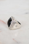 A silver signet ring with a large, faceted black oval stone, resting on a light marble surface.
