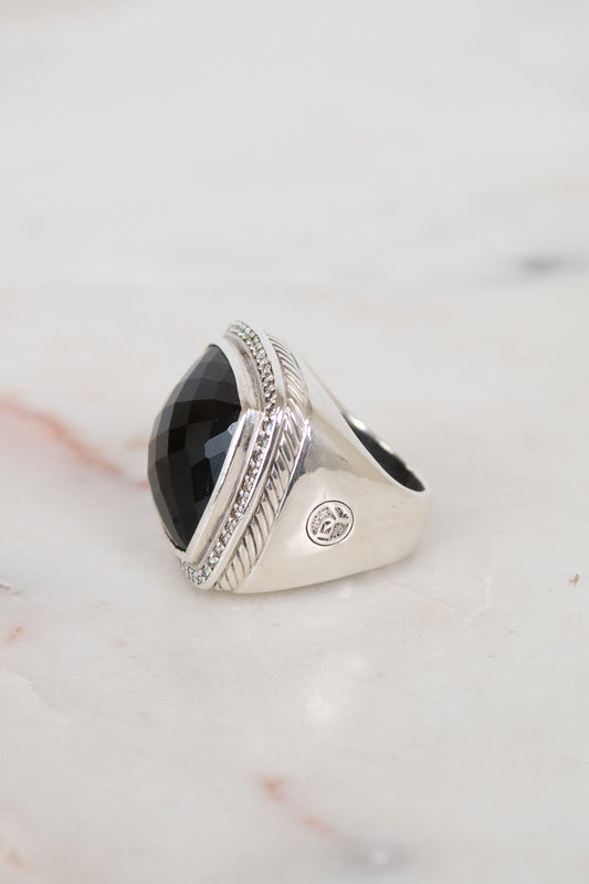 A silver signet ring with a large, faceted black oval stone, resting on a light marble surface.