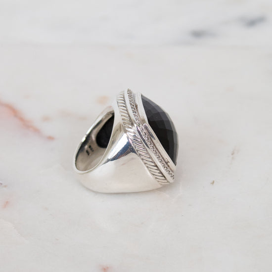 A silver signet-style ring with a large oval black stone, photographed on a light marble surface.