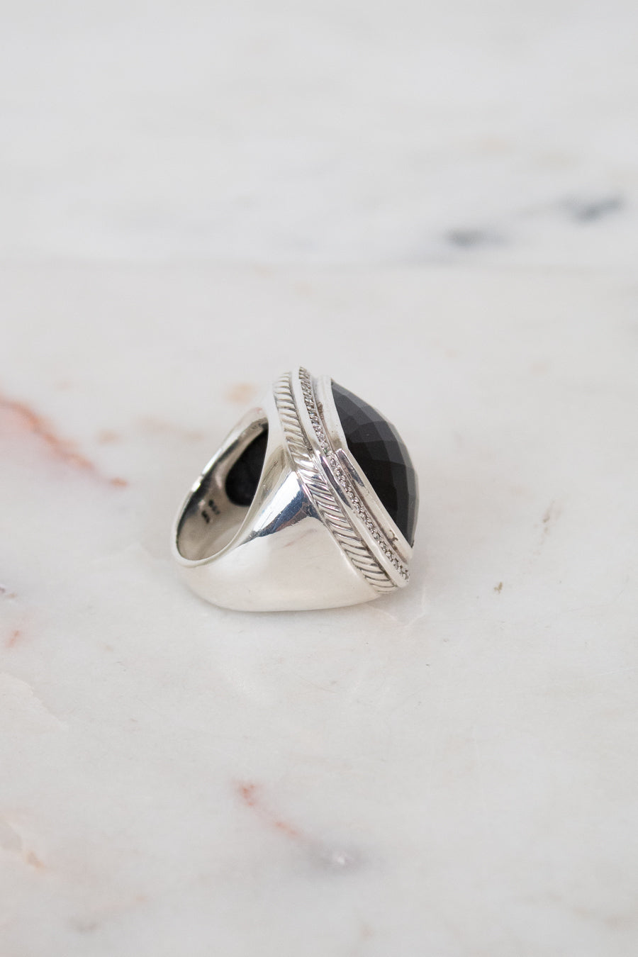 A silver signet-style ring with a large oval black stone, photographed on a light marble surface.