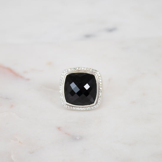 A square cushion-cut black gemstone ring with a silver-toned halo setting, displayed on a light marble surface.