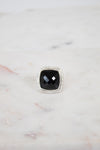 A square cushion-cut black gemstone ring with a silver-toned halo setting, displayed on a light marble surface.