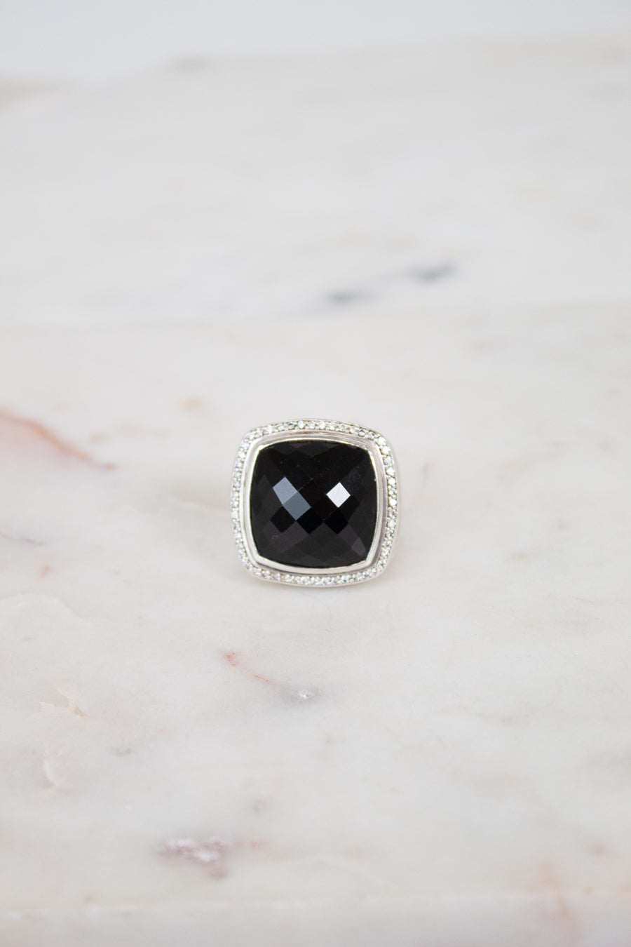 A square cushion-cut black gemstone ring with a silver-toned halo setting, displayed on a light marble surface.