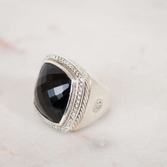 A silver signet ring with a large, faceted black onyx stone, accented with small clear stones along the top border, resting on a light marble surface.
