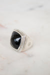 A silver signet ring with a large, faceted black onyx stone, accented with small clear stones along the top border, resting on a light marble surface.