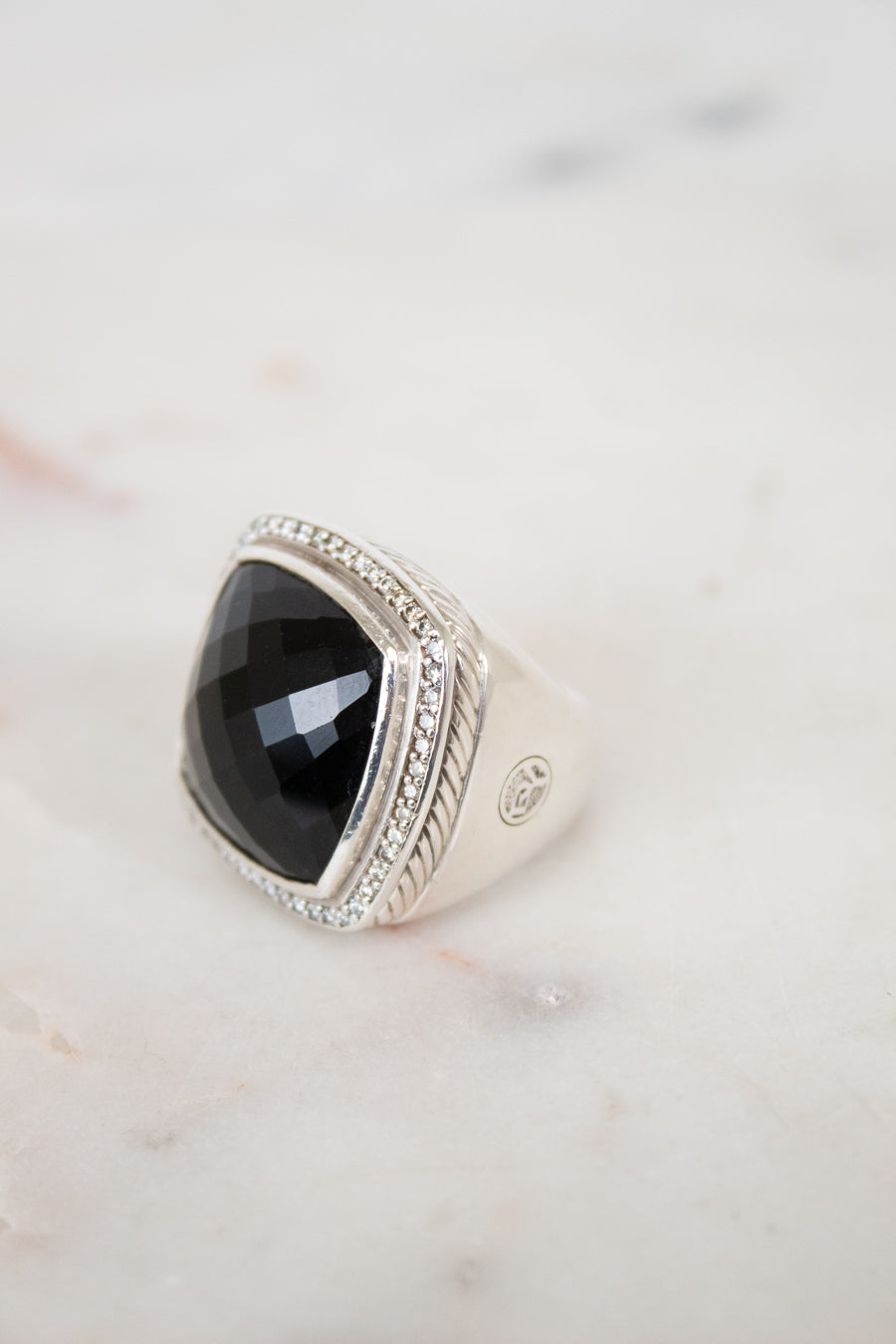 A silver signet ring with a large, faceted black onyx stone, accented with small clear stones along the top border, resting on a light marble surface.
