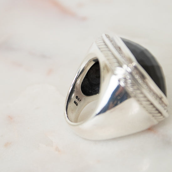 A silver signet-style ring with a large oval black stone, photographed close-up on a light marble surface.