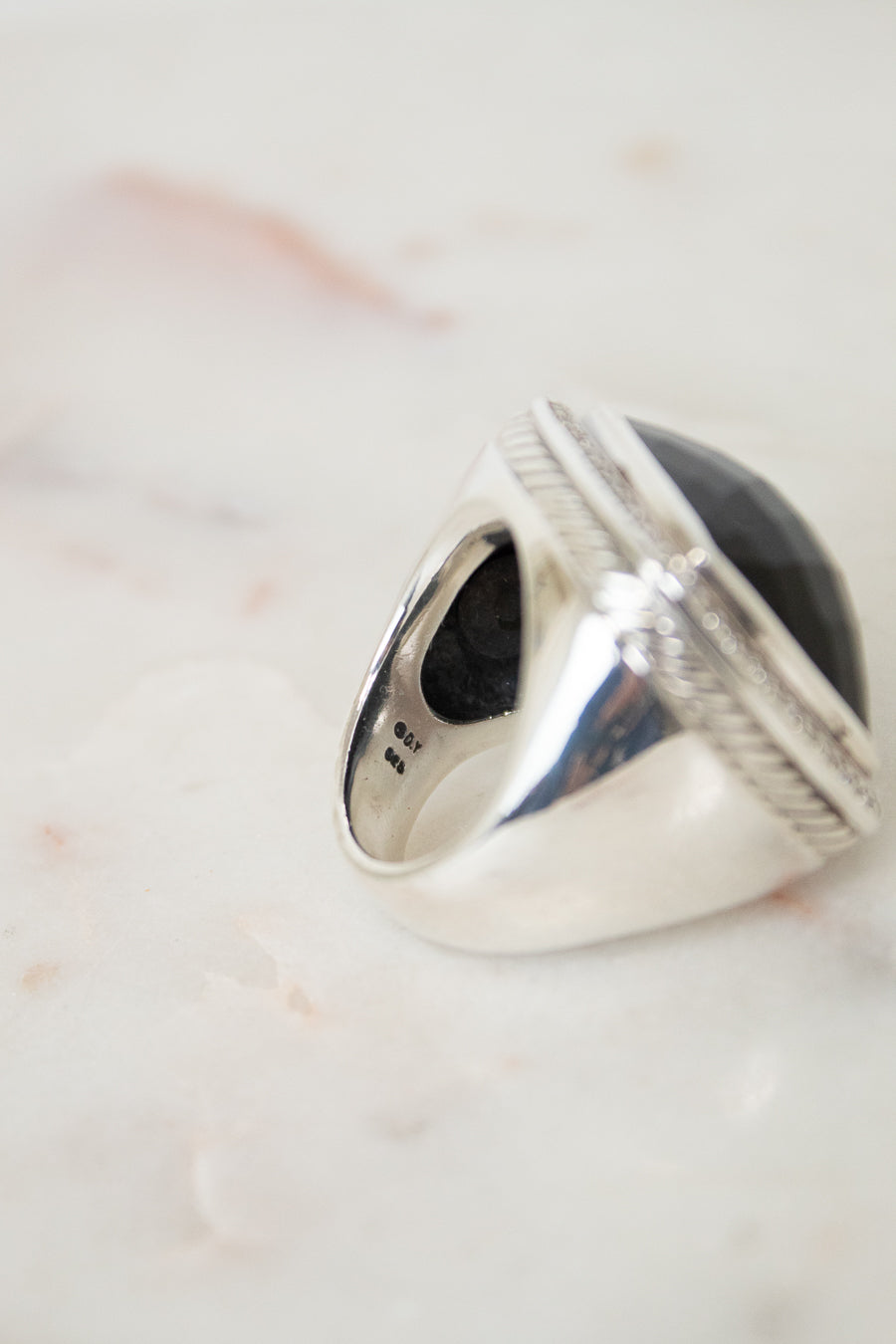 A silver signet-style ring with a large oval black stone, photographed close-up on a light marble surface.