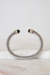 A twisted silver cable cuff bracelet with black onyx stone end caps, displayed on a white marble surface against a light gray background.