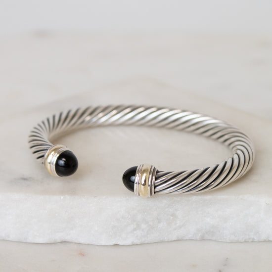 A silver cable-style cuff bracelet with black onyx bead ends and gold-toned accents, displayed on a white marble slab against a light neutral background.