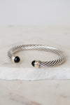 A silver cable-style cuff bracelet with black onyx bead ends and gold-toned accents, displayed on a white marble slab against a light neutral background.