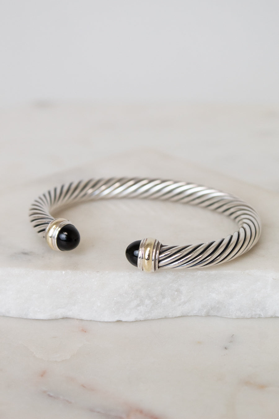 A silver cable-style cuff bracelet with black onyx bead ends and gold-toned accents, displayed on a white marble slab against a light neutral background.