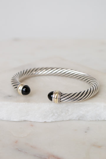 A silver cable-style cuff bracelet with black onyx bead ends and gold-toned accents, displayed on a white marble slab against a light neutral background.