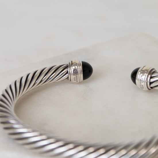 A silver cable-wrapped cuff bracelet with black onyx stone end caps, resting on a light marble surface.
