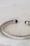 A silver cable-wrapped cuff bracelet with black onyx stone end caps, resting on a light marble surface.