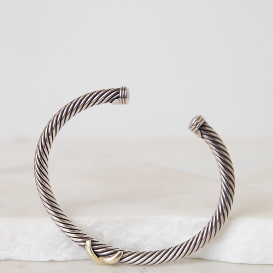 A silver-toned twisted cable cuff bracelet with a small gold accent at the front, displayed on a white marble slab against a light gray background.