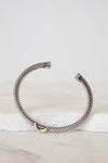 A silver-toned twisted cable cuff bracelet with a small gold accent at the front, displayed on a white marble slab against a light gray background.