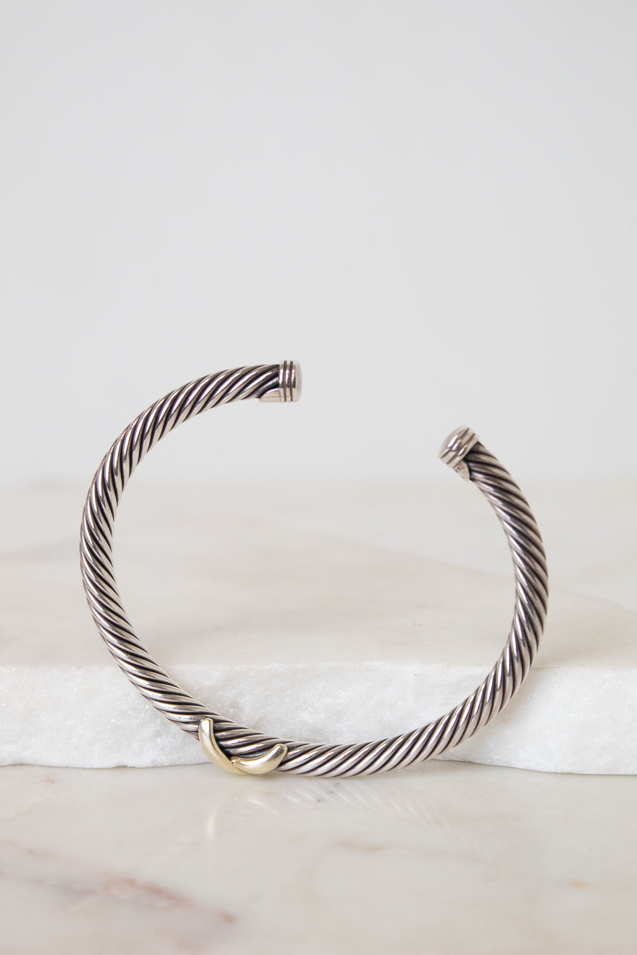 A silver-toned twisted cable cuff bracelet with a small gold accent at the front, displayed on a white marble slab against a light gray background.