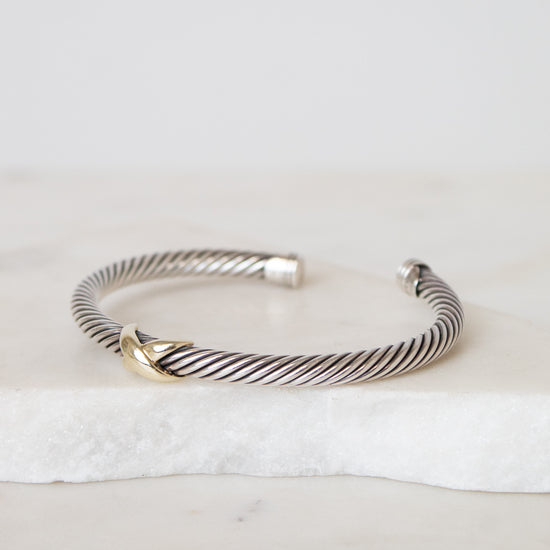 A silver cable-style cuff bracelet with a small gold accent at the front, displayed on a white marble slab against a light neutral background.