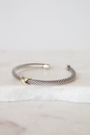 A silver cable-style cuff bracelet with a small gold accent at the front, displayed on a white marble slab against a light neutral background.