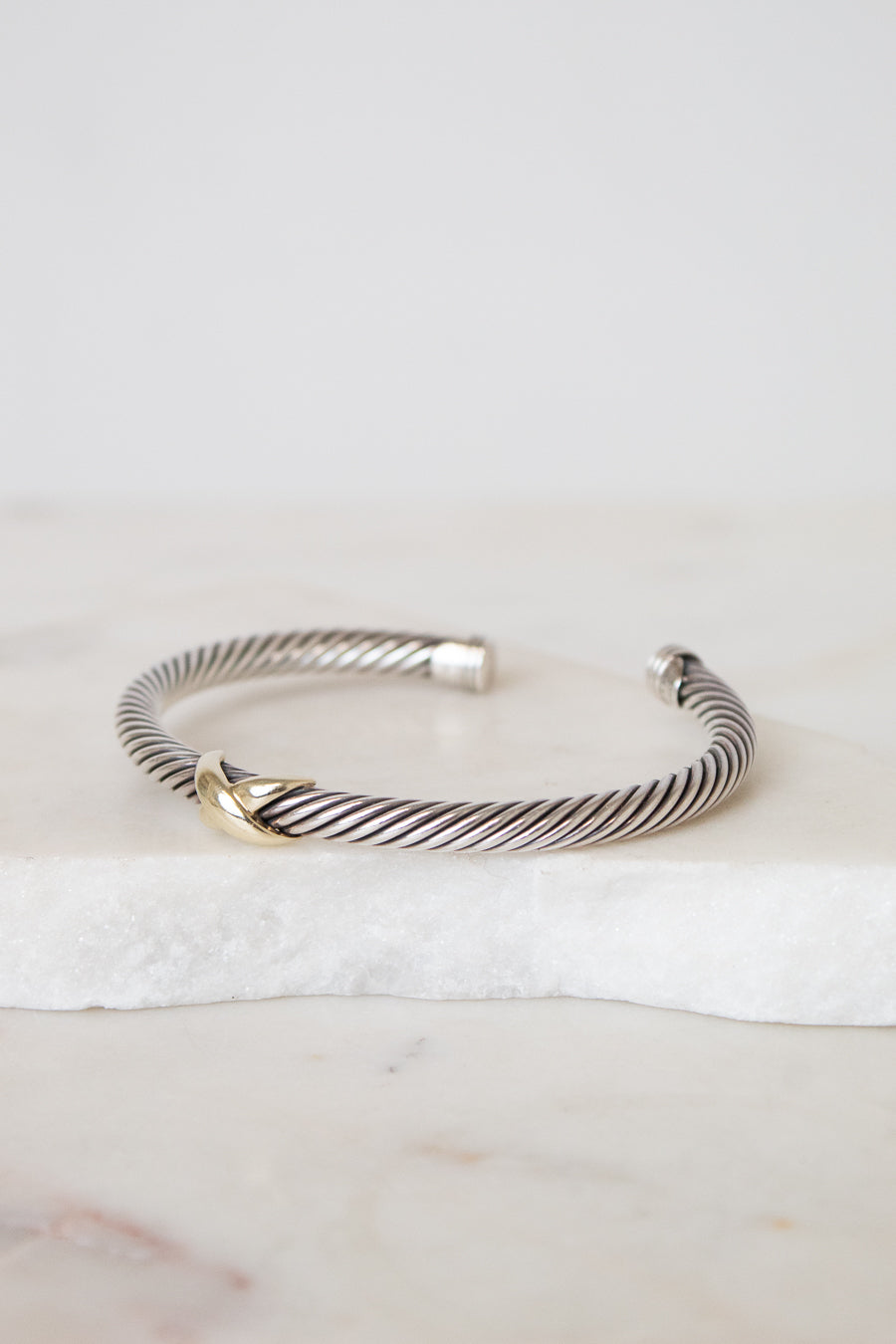 A silver cable-style cuff bracelet with a small gold accent at the front, displayed on a white marble slab against a light neutral background.