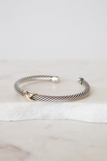 A silver cable-style cuff bracelet with a small gold accent at the front, displayed on a white marble slab against a light neutral background.