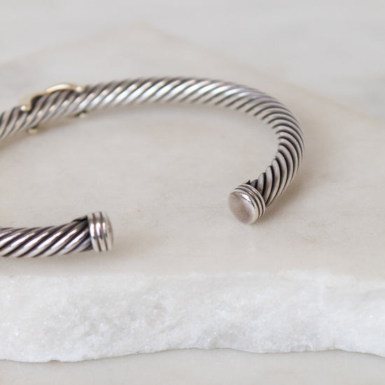 A silver cable-wrapped cuff bracelet with polished end caps, resting on a white marble slab.