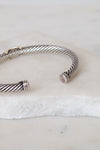 A silver cable-wrapped cuff bracelet with polished end caps, resting on a white marble slab.