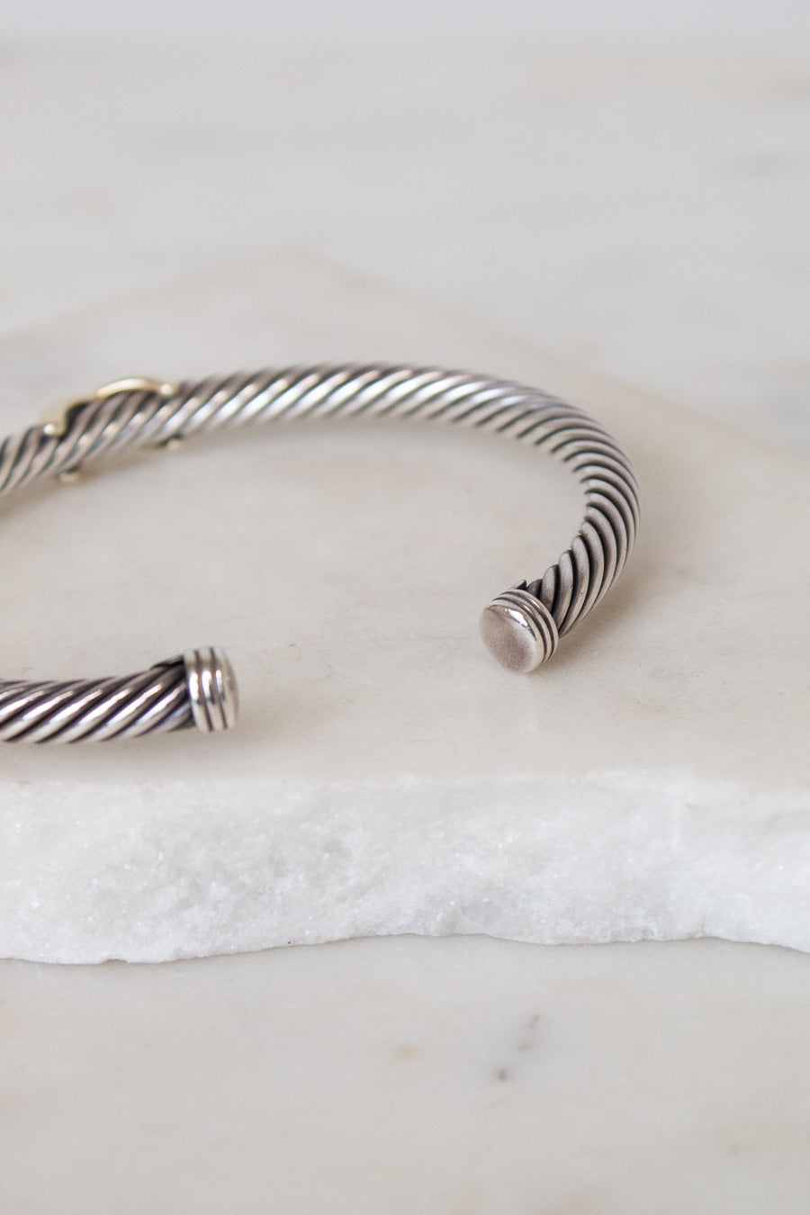 A silver cable-wrapped cuff bracelet with polished end caps, resting on a white marble slab.