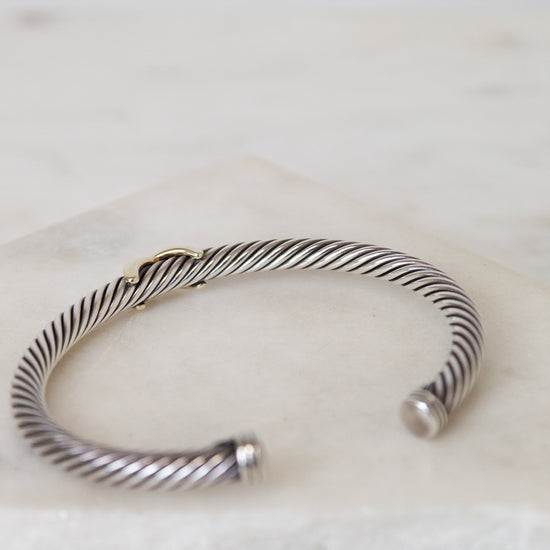 A silver cable-style cuff bracelet with a twisted wire pattern and a small gold accent, resting on a white marble surface.