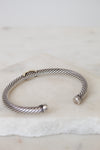 A silver cable-style cuff bracelet with a twisted wire pattern and a small gold accent, resting on a white marble surface.