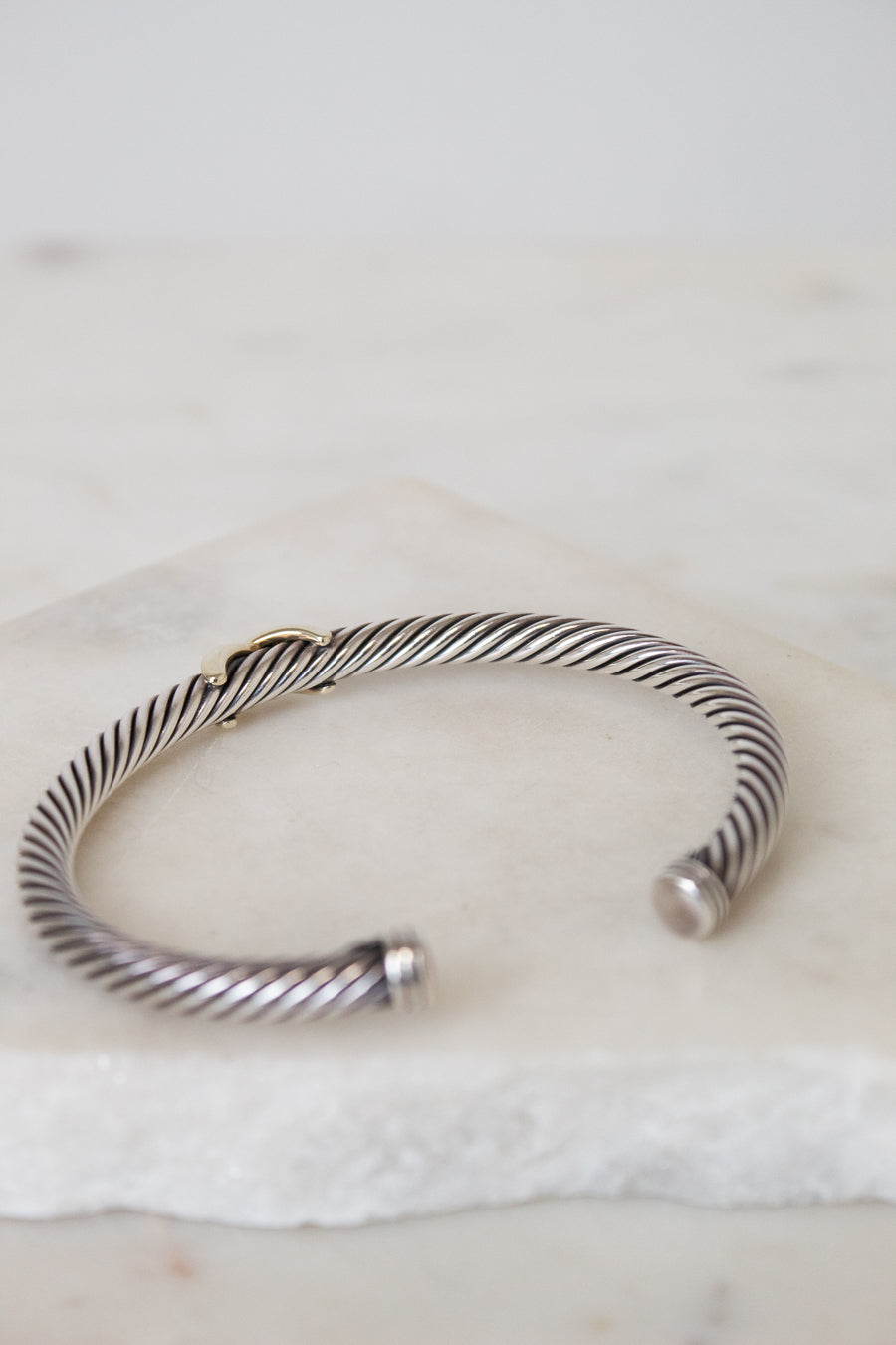 A silver cable-style cuff bracelet with a twisted wire pattern and a small gold accent, resting on a white marble surface.