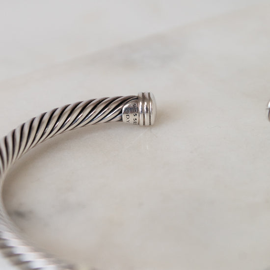 A close-up of a silver-toned twisted cable bangle bracelet resting on a light marble surface, with a small gold-toned accent visible at the front. 