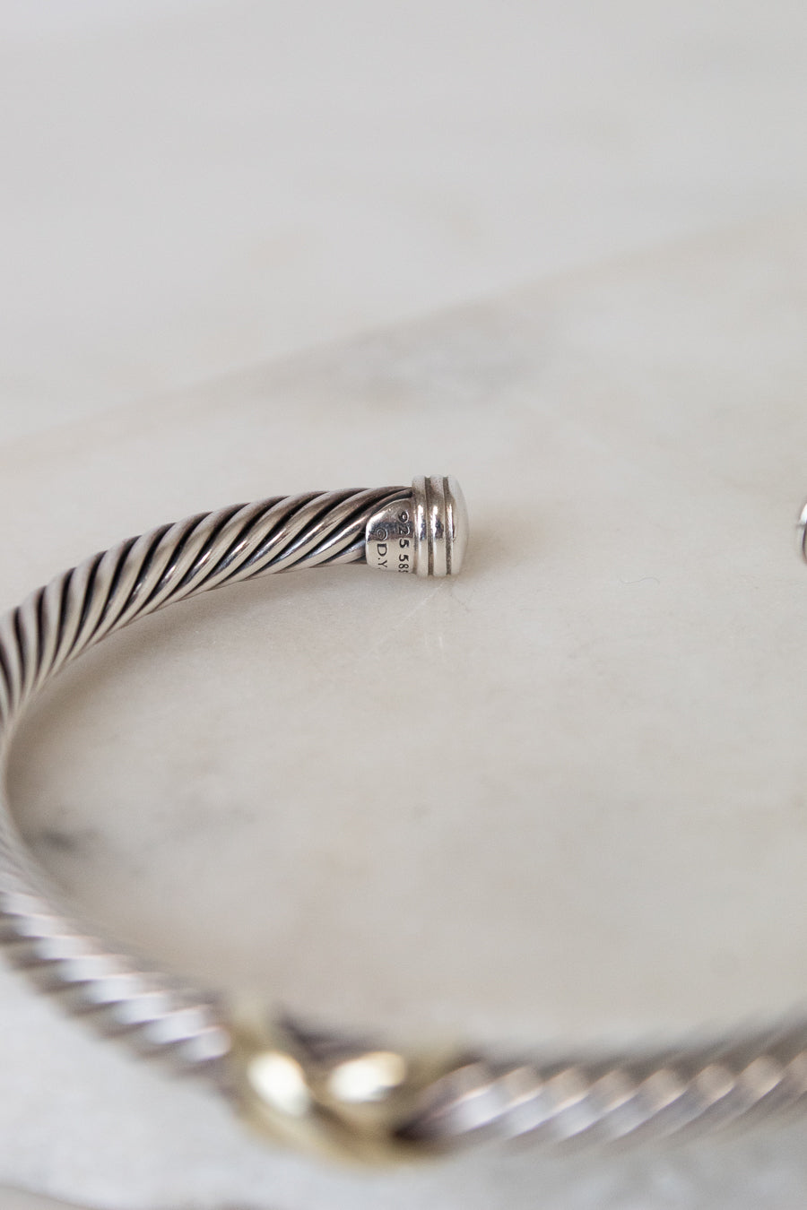 A close-up of a silver-toned twisted cable bangle bracelet resting on a light marble surface, with a small gold-toned accent visible at the front. 