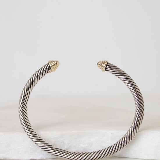 A twisted cable-style cuff bracelet in silver-tone metal with small gold-toned tips, displayed on a white marble surface against a light gray background.