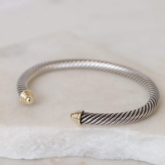 A silver-toned twisted cable open cuff bracelet with gold-toned end caps, resting on a white stone slab against a light neutral background.