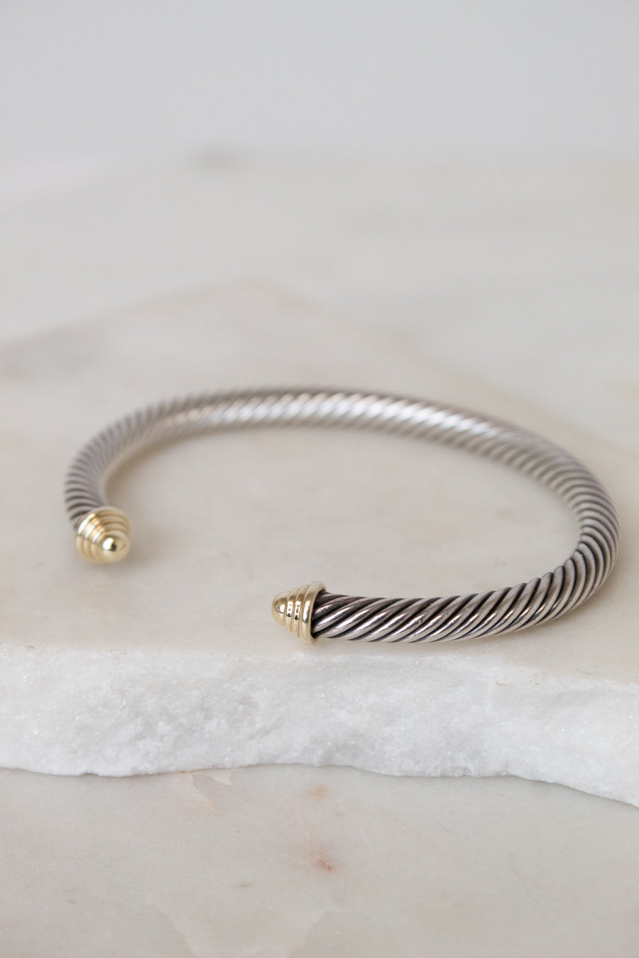 A silver-toned twisted cable open cuff bracelet with gold-toned end caps, resting on a white stone slab against a light neutral background.
