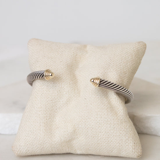 A silver twisted cable cuff bracelet with gold details displayed on a small beige fabric jewelry pillow over a white marble surface.