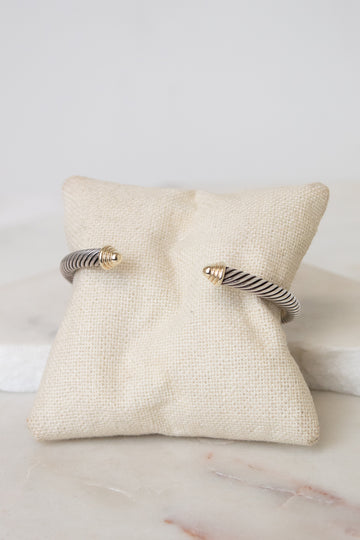 A silver twisted cable cuff bracelet with gold details displayed on a small beige fabric jewelry pillow over a white marble surface.