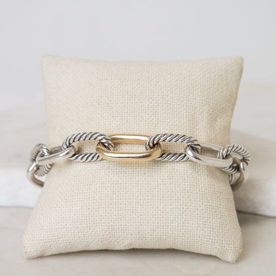 A gold and silver-toned cable link chain bracelet displayed across a cream fabric jewelry pillow on a light marble surface.