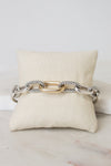 A gold and silver-toned cable link chain bracelet displayed across a cream fabric jewelry pillow on a light marble surface.