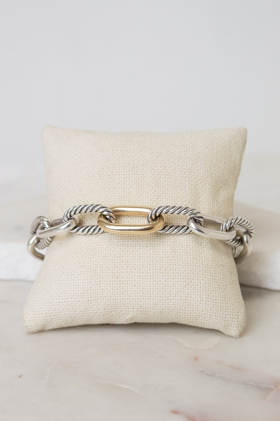 A gold and silver-toned cable link chain bracelet displayed across a cream fabric jewelry pillow on a light marble surface.