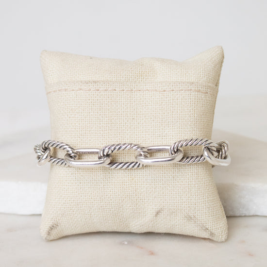 A silver-toned cable link chain bracelet displayed across a cream fabric jewelry pillow on a light marble surface.