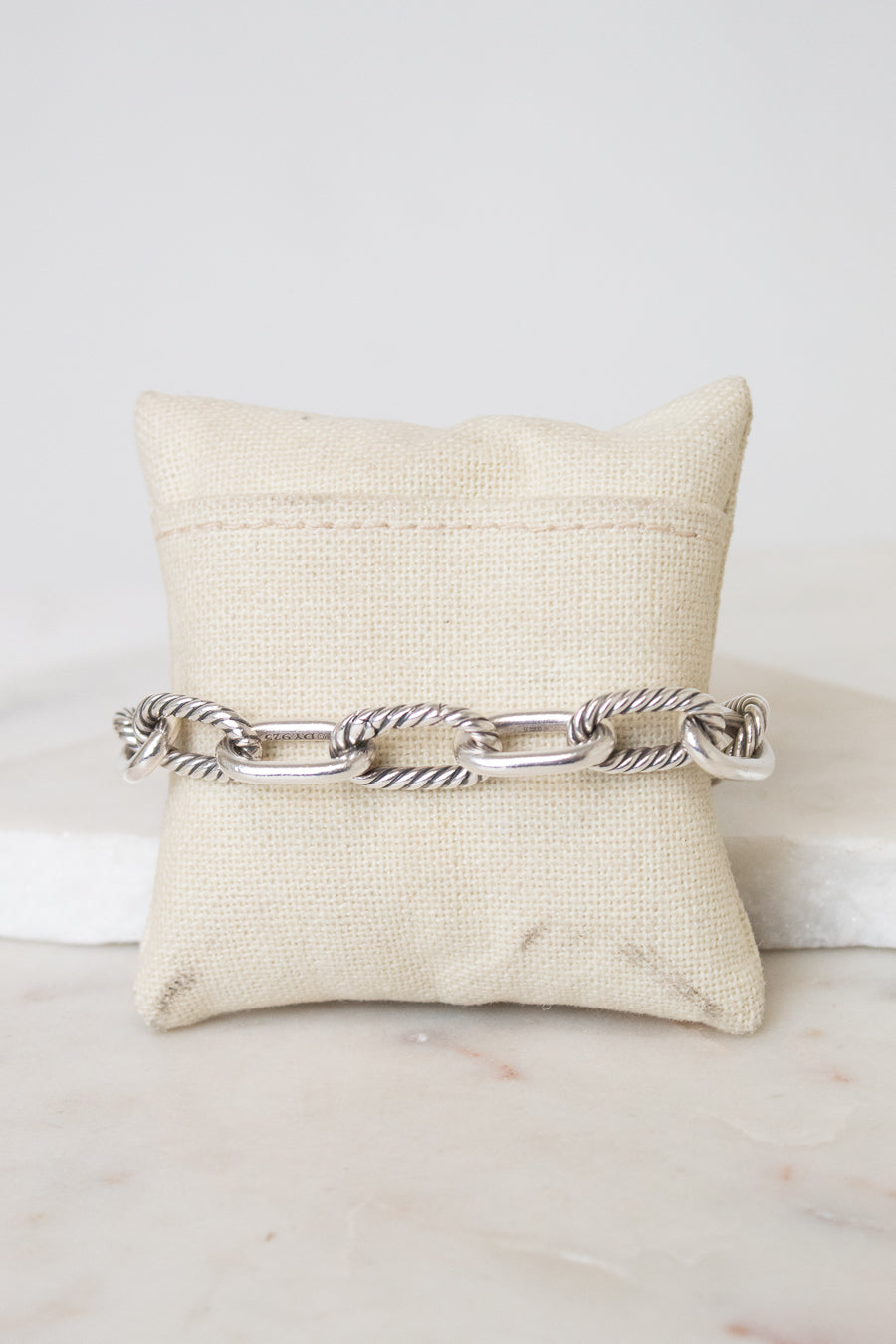 A silver-toned cable link chain bracelet displayed across a cream fabric jewelry pillow on a light marble surface.