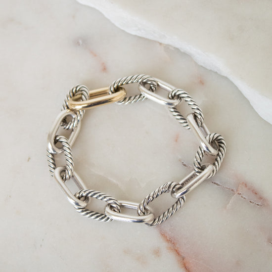 A silver cable-link chain bracelet with a gold clasp, displayed on a light beige marble surface.