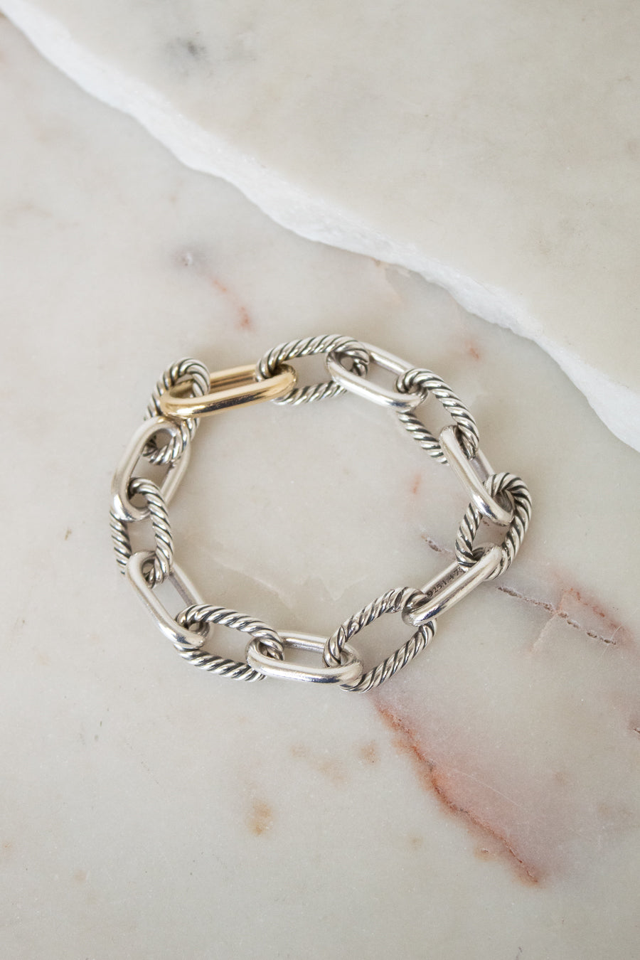 A silver cable-link chain bracelet with a gold clasp, displayed on a light beige marble surface.